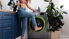 Žena v kuchyni otvára práčku nohou, drží bielizeň pripravenú na pranie.