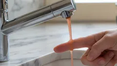 Oranžová voda tečúca z kuchynského kohútika, osoba kontroluje prstom kvalitu vody.