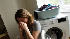 Žena sedí pred práčkou a drží si tvár, vedľa je koš s bielizňou.