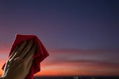 Ruka čistí okno handrou pri západe slnka, večerná obloha v pozadí.